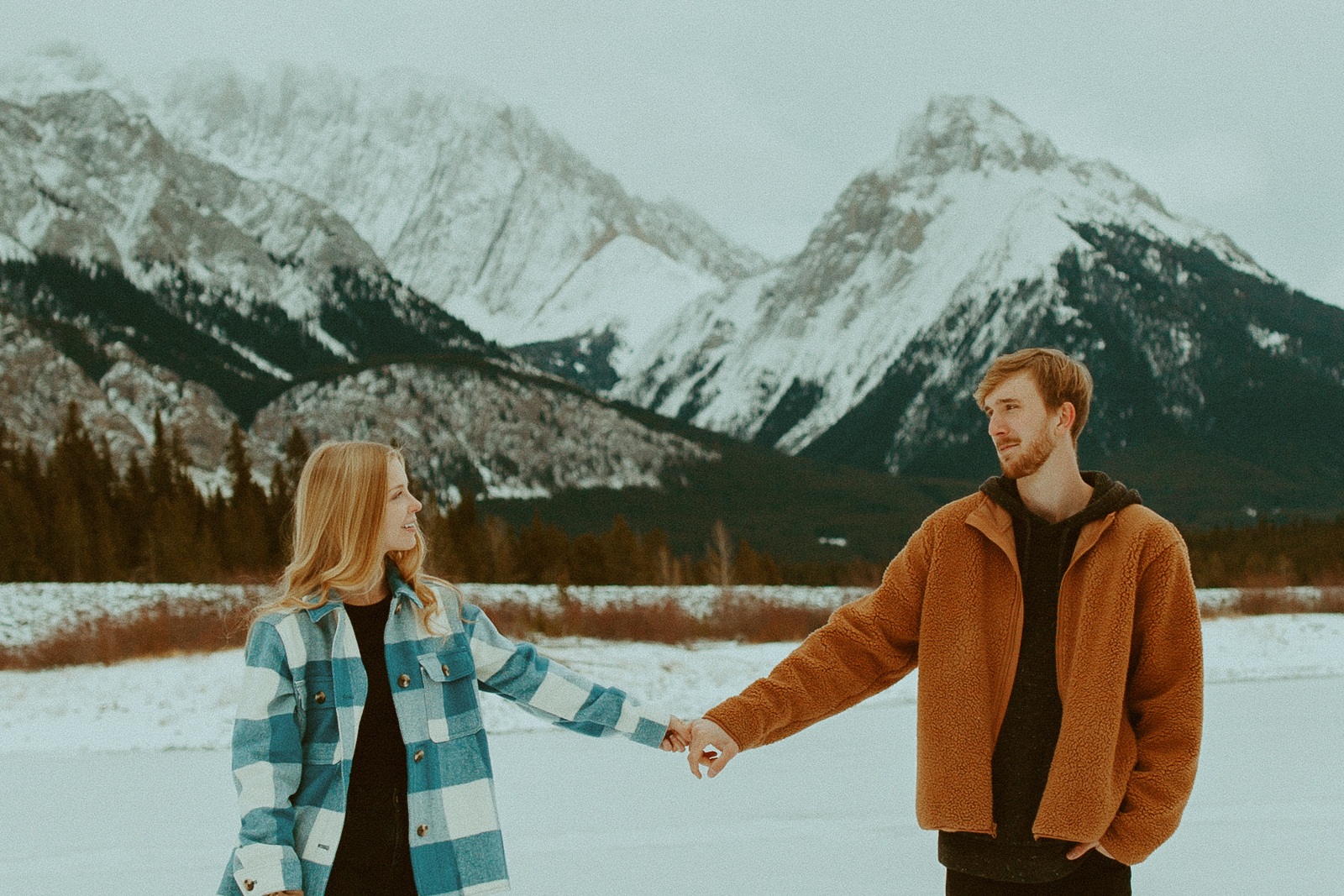 couple holds hands, standing in front of mountains along the shore of lower Kananaskis lake